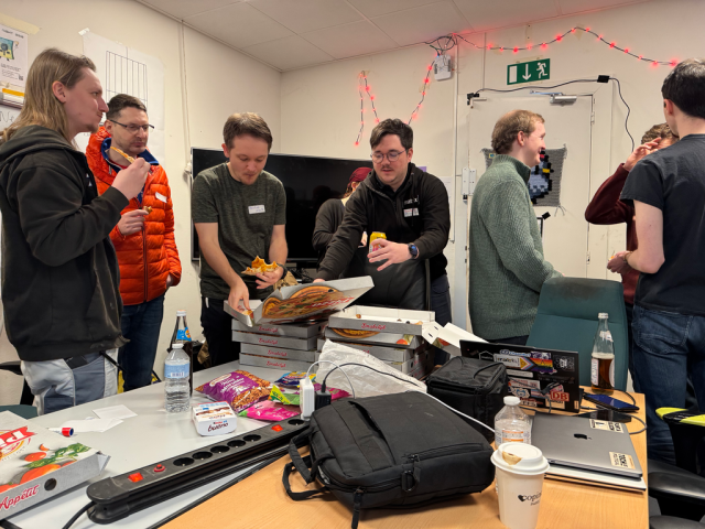 A picture of people gathering around a stack of pizzas and talking.