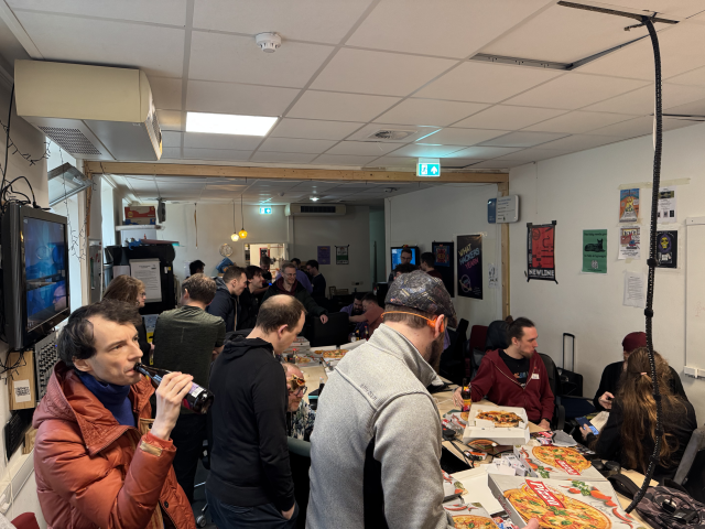 A picture of people eating and drinking, gathered around tables. On the tables there are stacks of pizzas.