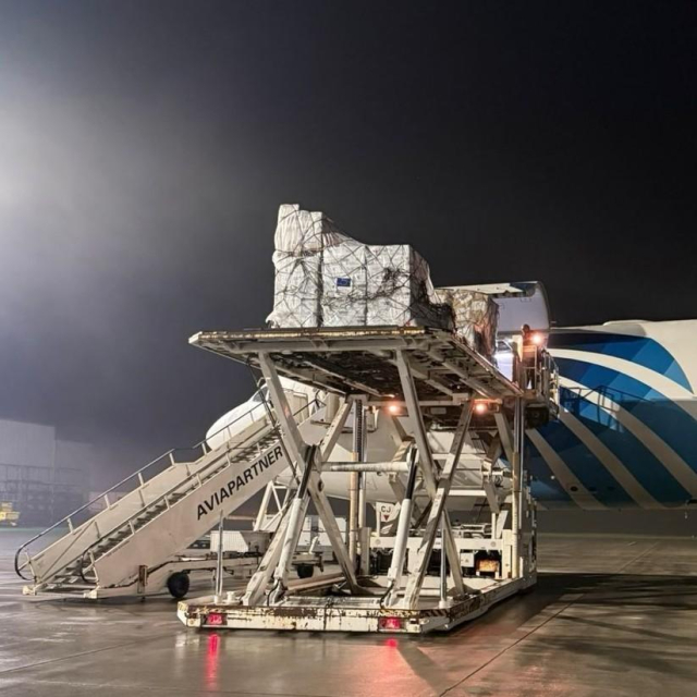 A cargo pallet loaded with supplies is being lifted onto an airplane at night. The pallet is secured with netting, and the scene is illuminated by airport lighting. The aircraft has a blue and white design, and a mobile staircase is positioned beside it. The background is dark, and the tarmac is wet, reflecting the lights.  