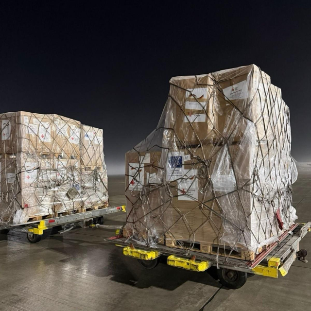 Two large pallets of humanitarian supplies are loaded onto airport trolleys on a tarmac at night. The pallets are wrapped in plastic and secured with netting, with labels indicating they contain Humanitarian aid supplies. The scene is dimly lit, with a dark sky overhead and no other visible people or aircraft nearby.  