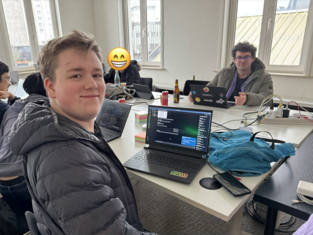 A picture of a person proudly standing next to their laptop. Their laptop has a the new client they work on open, The client now supports displaying the room list and intentional mentions.

In the background, someone smiles as they work on their own laptop.