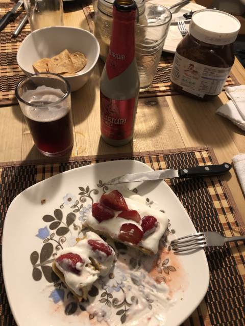 A waffle swimming in whipped cream and sliced strawberries, and a glass of fruit beer.