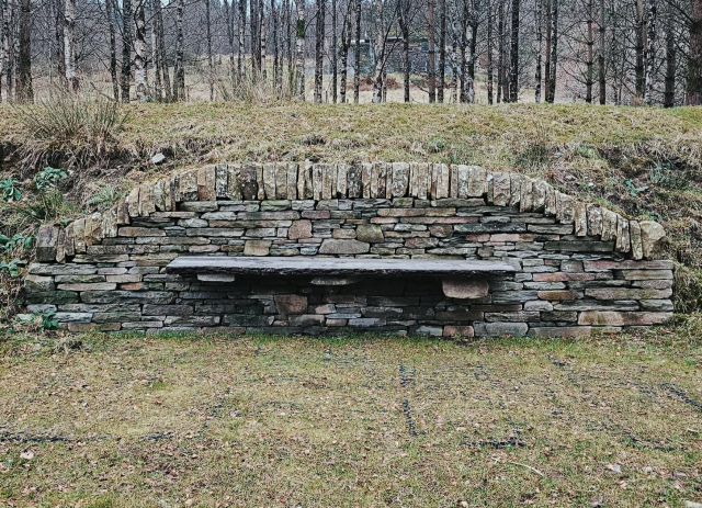 Wide view of a drystone bench set into a curved stone wall on a grassy bank beside Loch Voil. The wall is built from mixed local stone with upright coping stones along the top. Leafless trees and a sloping hillside rise behind the bench.