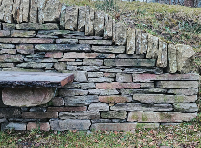 Close-up of a drystone bench built into a low stone wall. The stones are layered in horizontal courses with upright coping stones on top. A thick wooden slab forms the seat, supported by projecting stones beneath. Grass and moss surround the base.