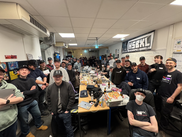 A wide-angle picture of a completely packed room. People are around a long table with plenty of pizza boxes and laptops. Everyone wears a Matrix cap and is smiling.