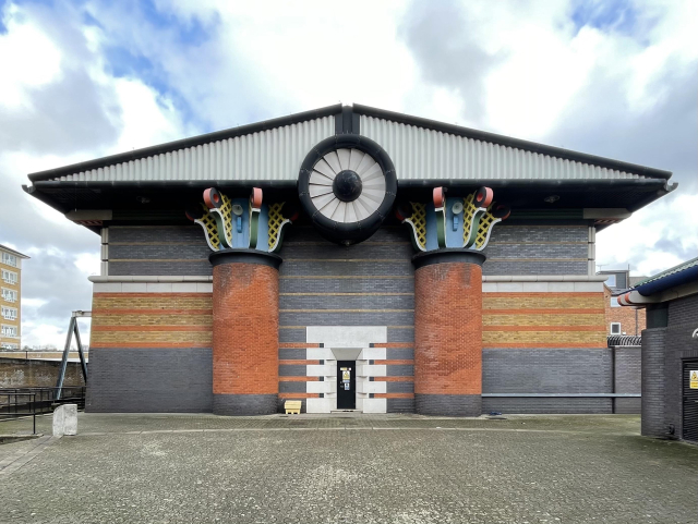 A building with grey and brown brick walls supporting a black and white pediment with a huge white fan in the centre. Two thick red brick columns are topped with blue, yellow, and red fins. The building complexly dwarfs its normal-sized door.