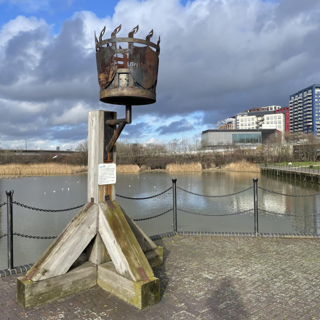 A large metal basket decorated with small flames and the British Gas logo, attached to a chunky wooden support. In the background is a former dock with reeds growing around the edges.