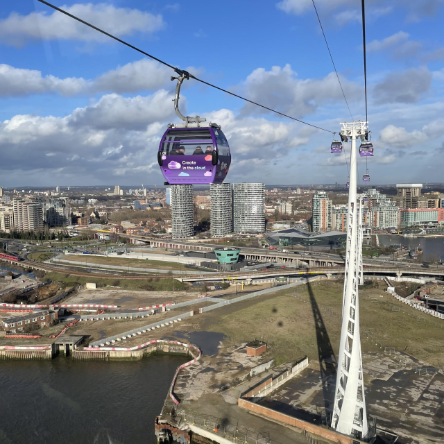View from a cable car. Another cable car is visible nearby, with more in the distance. A huge white spiral pylon is on the edge of the photo, standing in empty land. Part of the River Thames is visible in one corner.