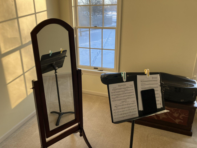 In the bottom right there is a music stand with an iPad and sheet music on it. In front of that is a full length mirror. The back of the music stand is reflected on that. 

Behind the mirror is the golden light of a sunset streaming in from the south facing window. To the right of the mirror is a window with a snowy front yard. 

And finally to right of that is a black cello case sitting on top off a brown chest.