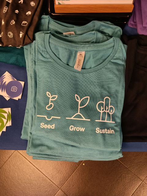 Blue T-shirt in table with other swag. Text it says "Seed, Grow, Sustain"