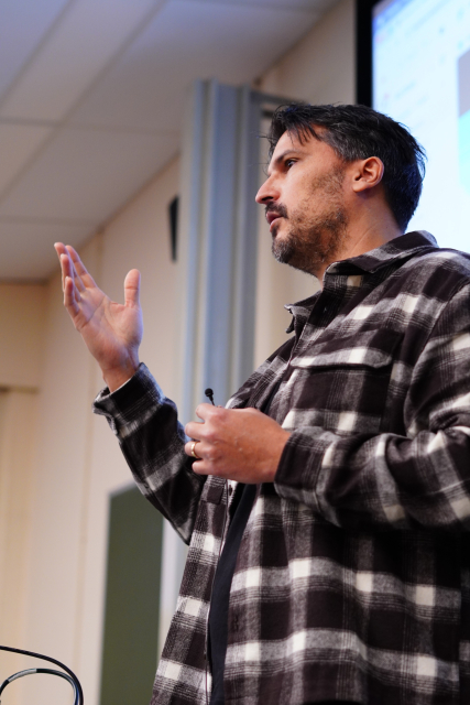 A portrait of Valerie talking to the audience. He is holding a microphone in one hand and gesturing with his second hand