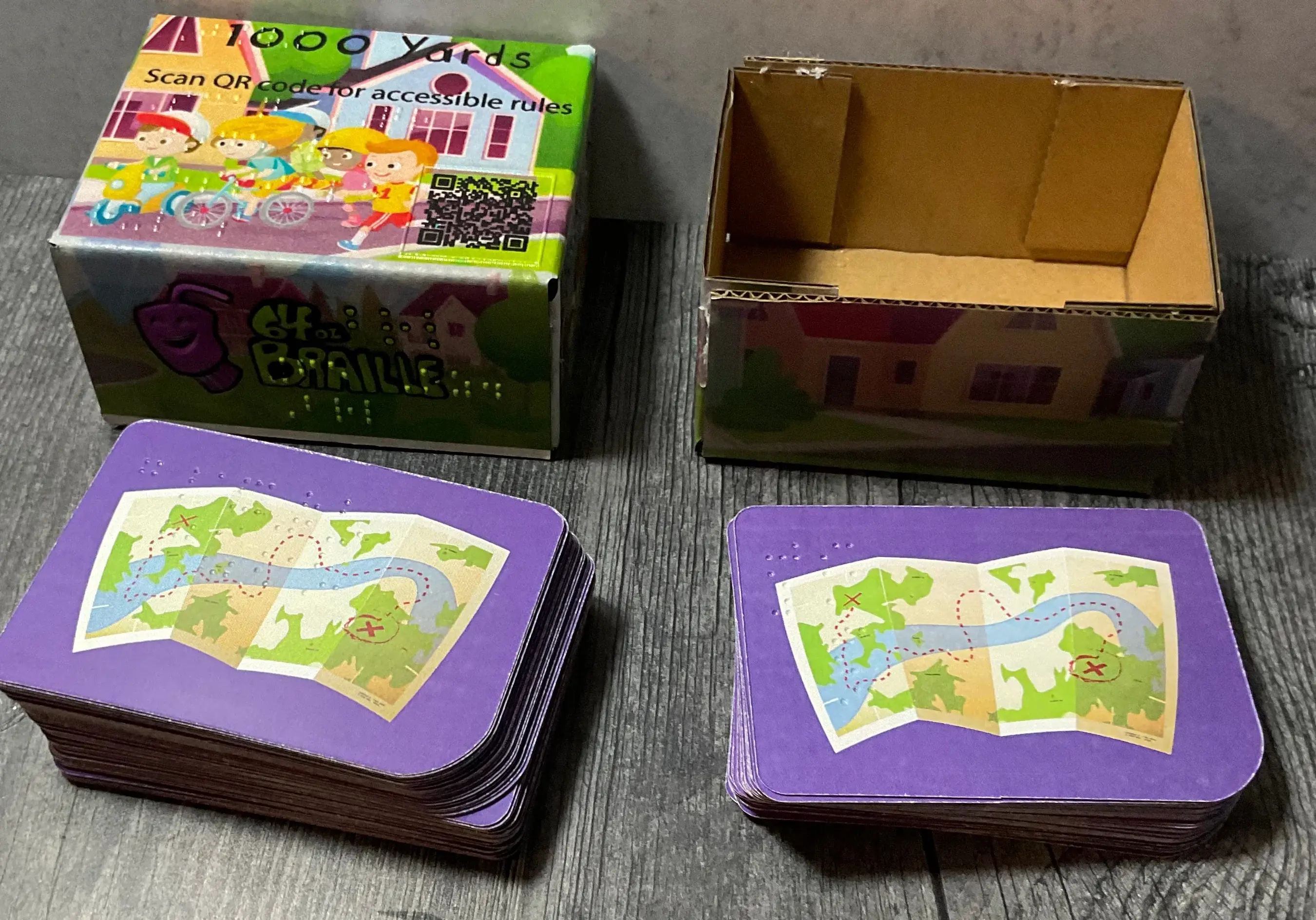 an unboxed card game called 1000 yards. There are 2 stacks of playing cards visible with Braile on them and the box has print, Braille, and a QR code visible.