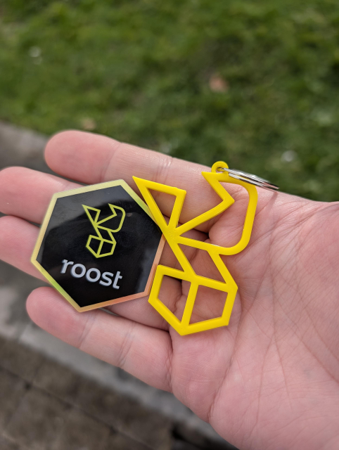 Holding a hexagonal sticker with the ROOST logo next to a yellow keychain of the same logo