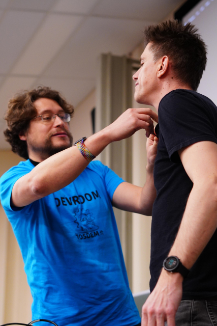 A portrait of Matthew attaching a mic to Neil as he prepares for his talk in the devroom.