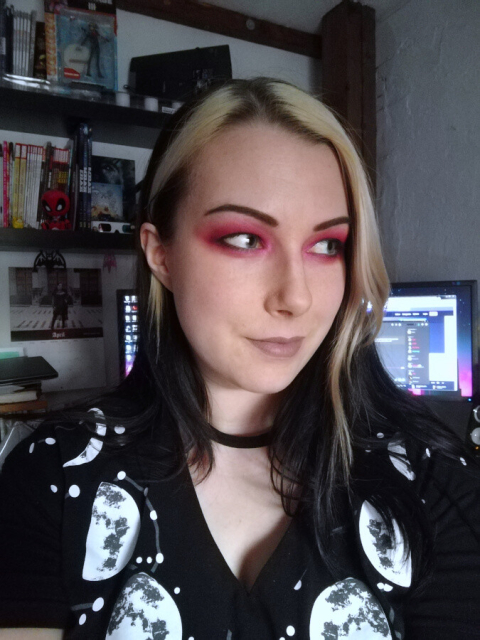 Selfie of myself in front of my computer. Looking sideways away from the viewer. Long black hair with blond streaks in front. Pink eye make up and nude lipstick. Wearing a black v-neck shirt with moon phases printed on. In the shelf in the background you can see comics, figures and other geeky objects. A "Goths for Sophie" calender is on the wall.