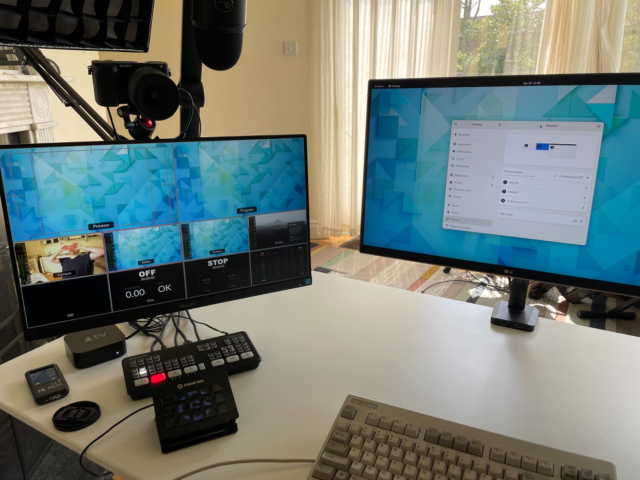 Photo of my white desk with two monitors, one running a video switcher (Atem) multiview with camera, slides, screen, and AirPlay views visible. The other running a GNOME 42 desktop. A white keyboard is partially visible too.