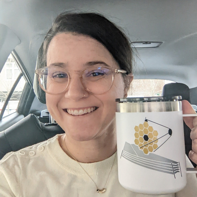 A selfie of Erin (brown hair pulled back in a bun, hazel eyes, large clear glasses, noise ring) smiling while holding a travel mug she made featuring the JW Space Telescope