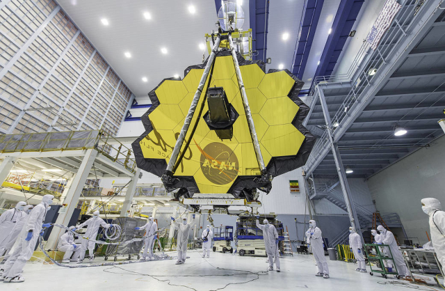 A picture of JWST in the clean room with people wearing clean suits surrounding it. The NASA logo is reflected in the gold mirror