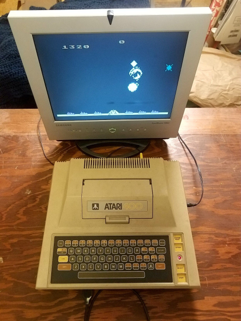 An Atari 400 computer connected to a 15-inch LCD TV displaying the game missile command. All are sitting on a wooden workbench.